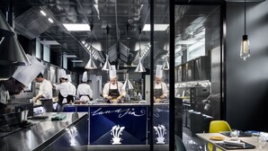 A modern kitchen with chefs preparing food, stainless steel appliances, and a blue counter with a logo. - Hotel Plaza e de Russie (Viareggio)