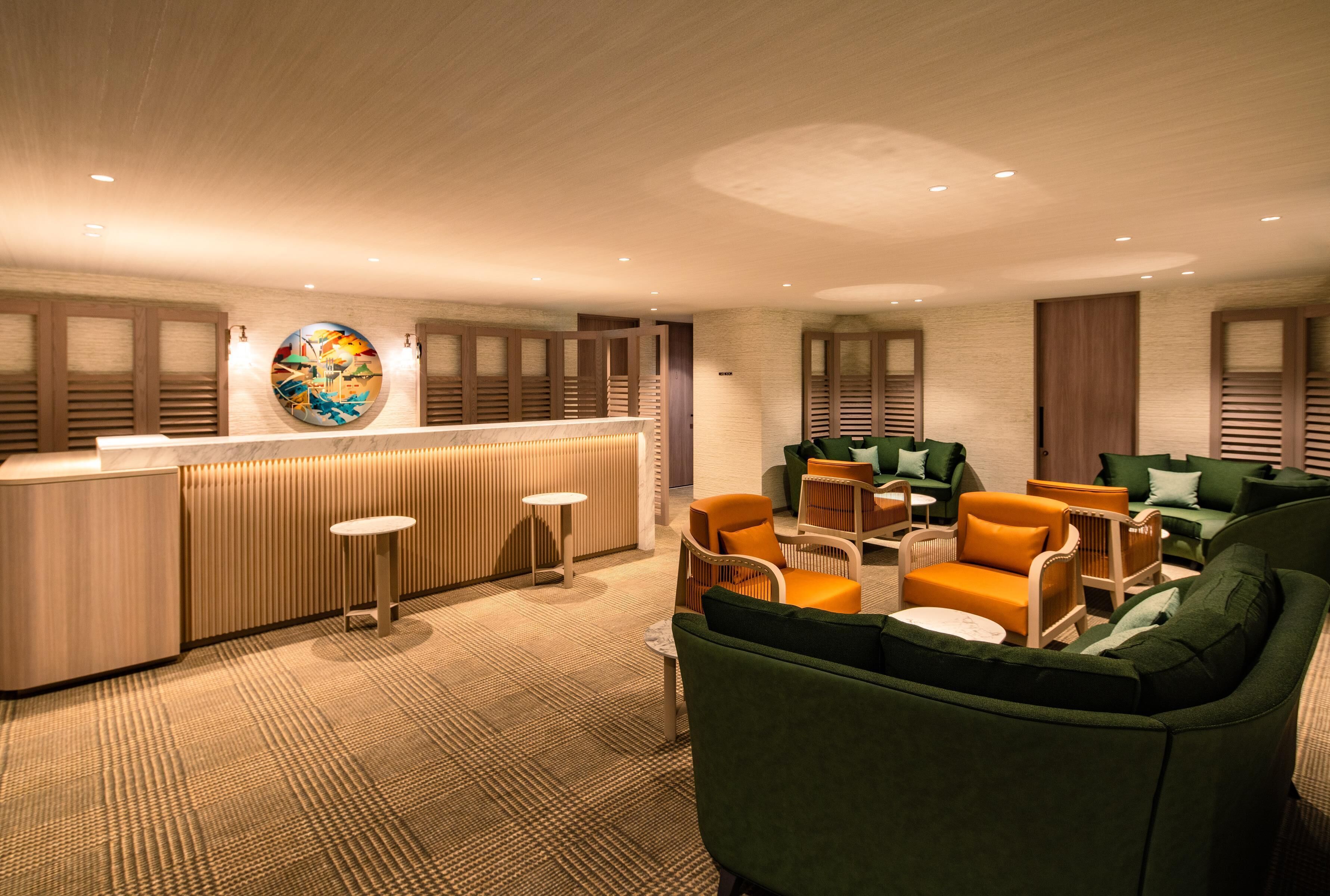 A modern lounge area with a wooden reception desk, green and orange seating, and a patterned carpet.