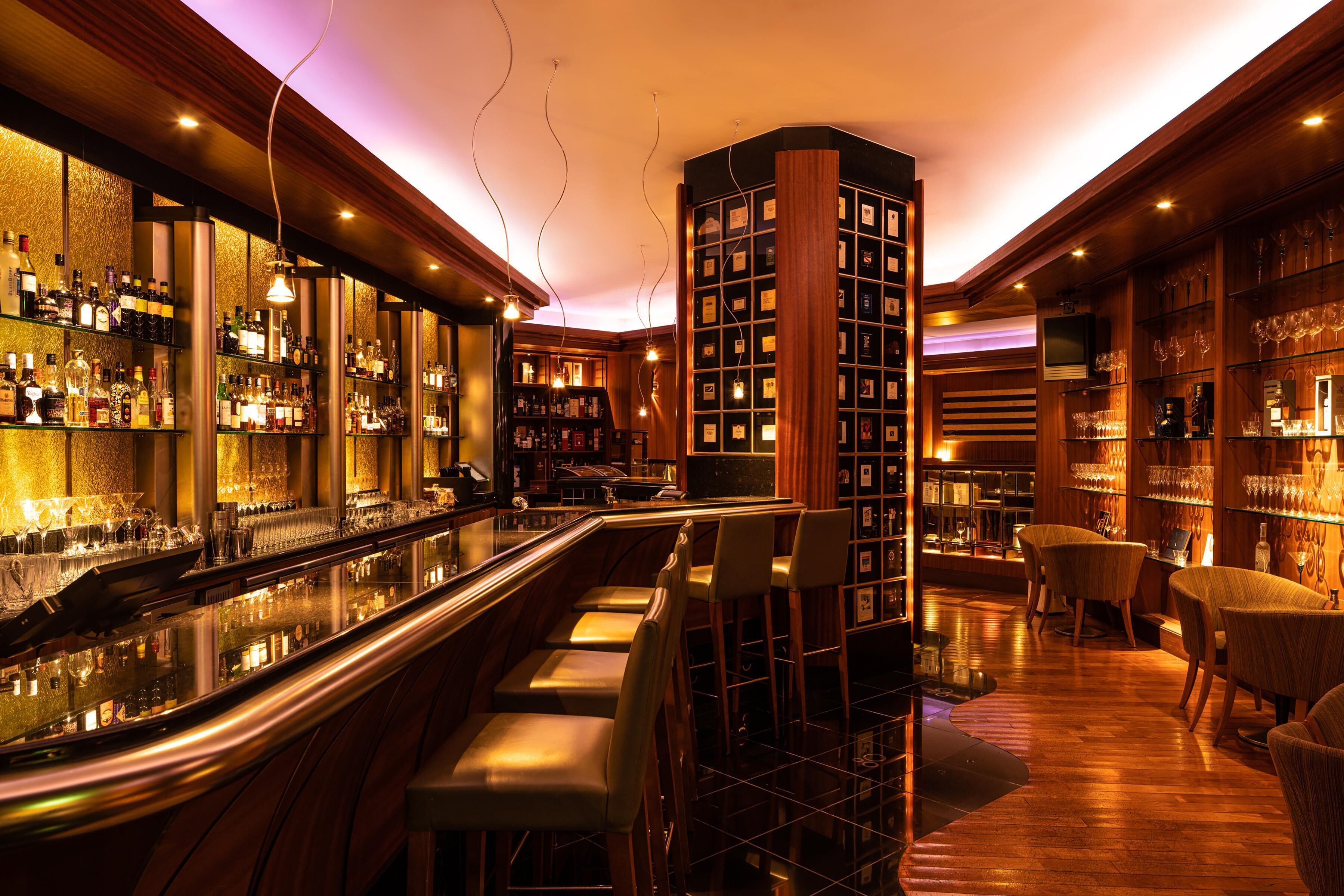 A bar area with a long counter, multiple stools, and shelves stocked with bottles and glasses.