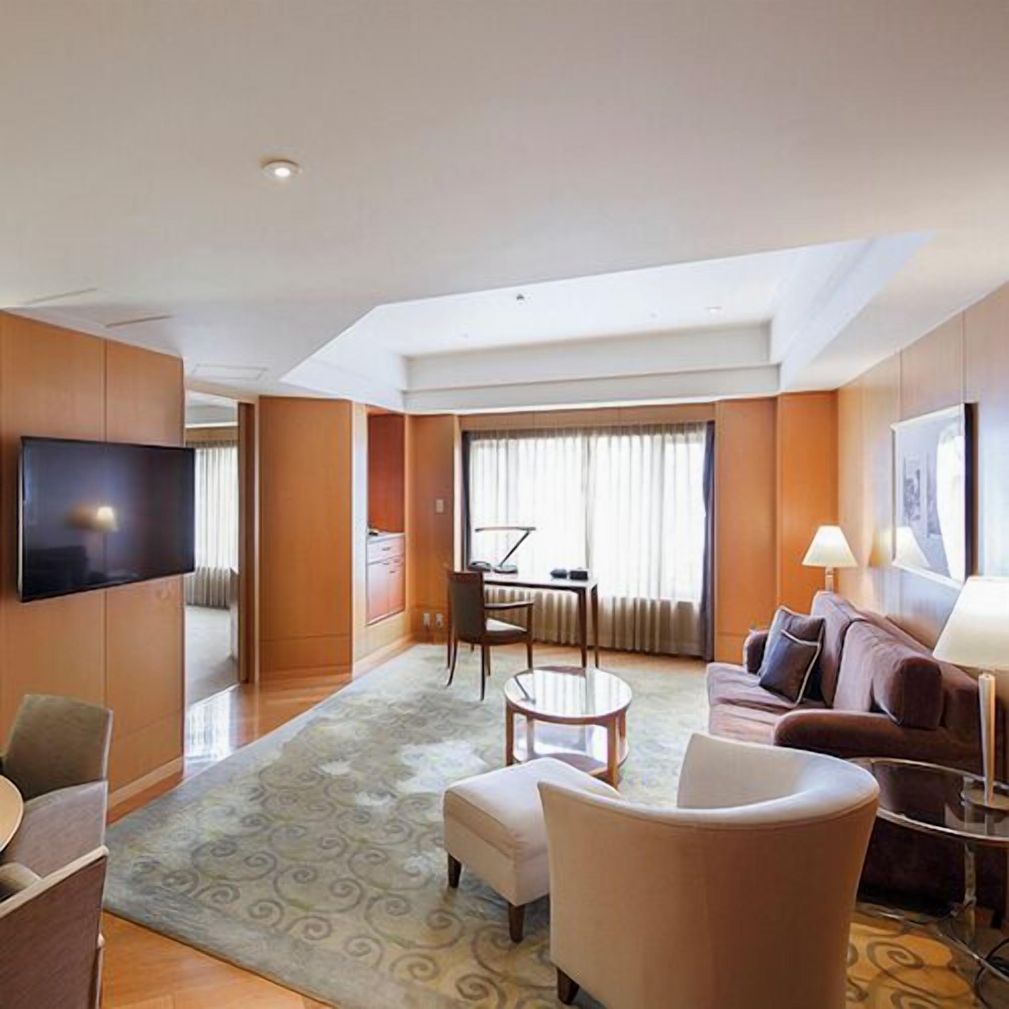 A hotel room with a flat-screen TV, a sofa, a dining table with chairs, a small round table, and a large window with curtains.