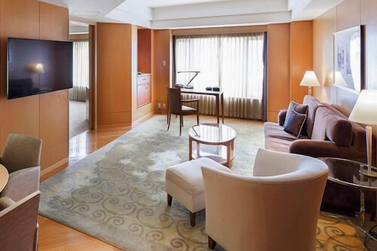 A hotel room with a flat-screen TV, a sofa, a dining table with chairs, a small round table, and a large window with curtains.