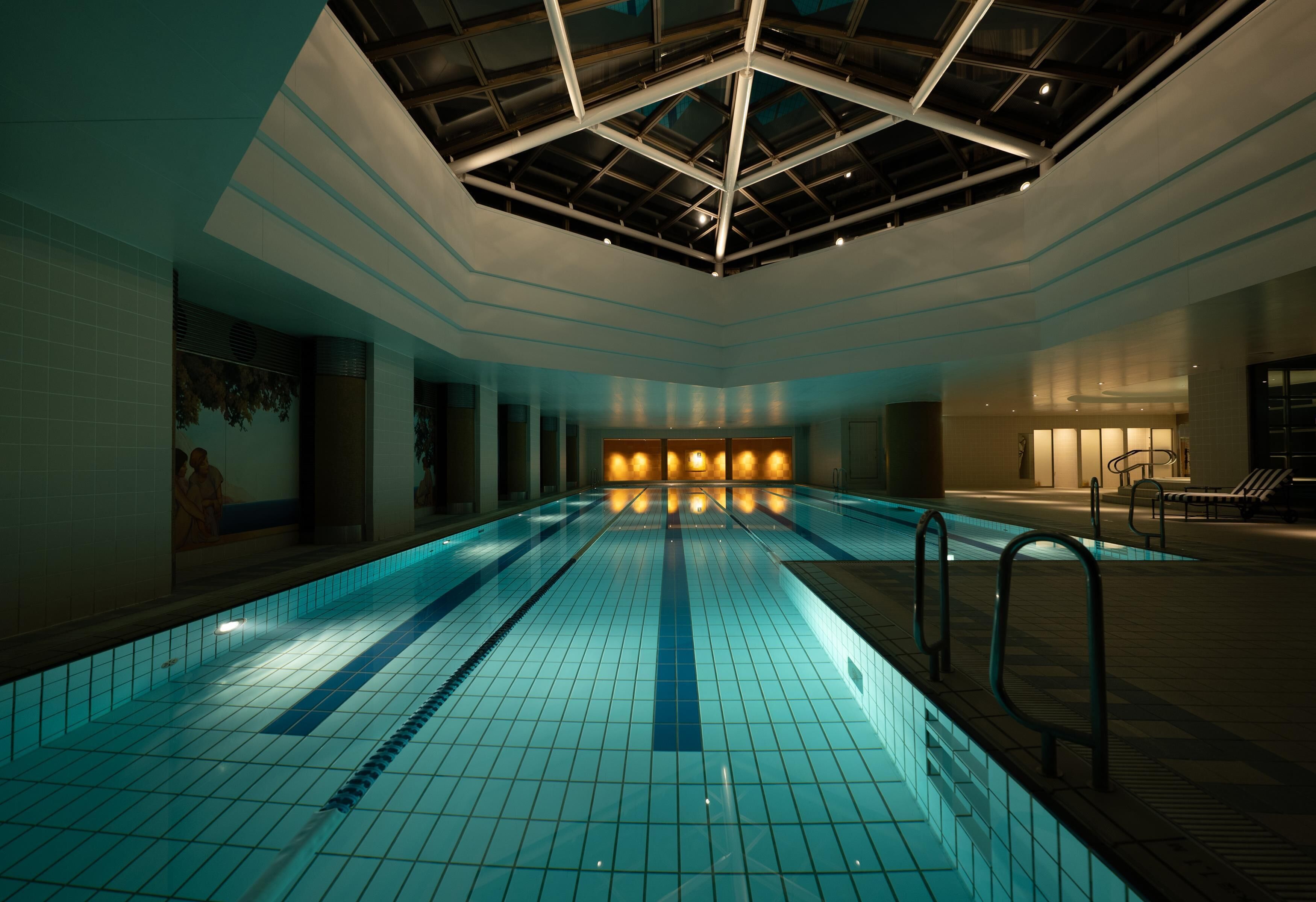 An indoor swimming pool with a geometric ceiling design, tiled floor, and a seating area with chairs.