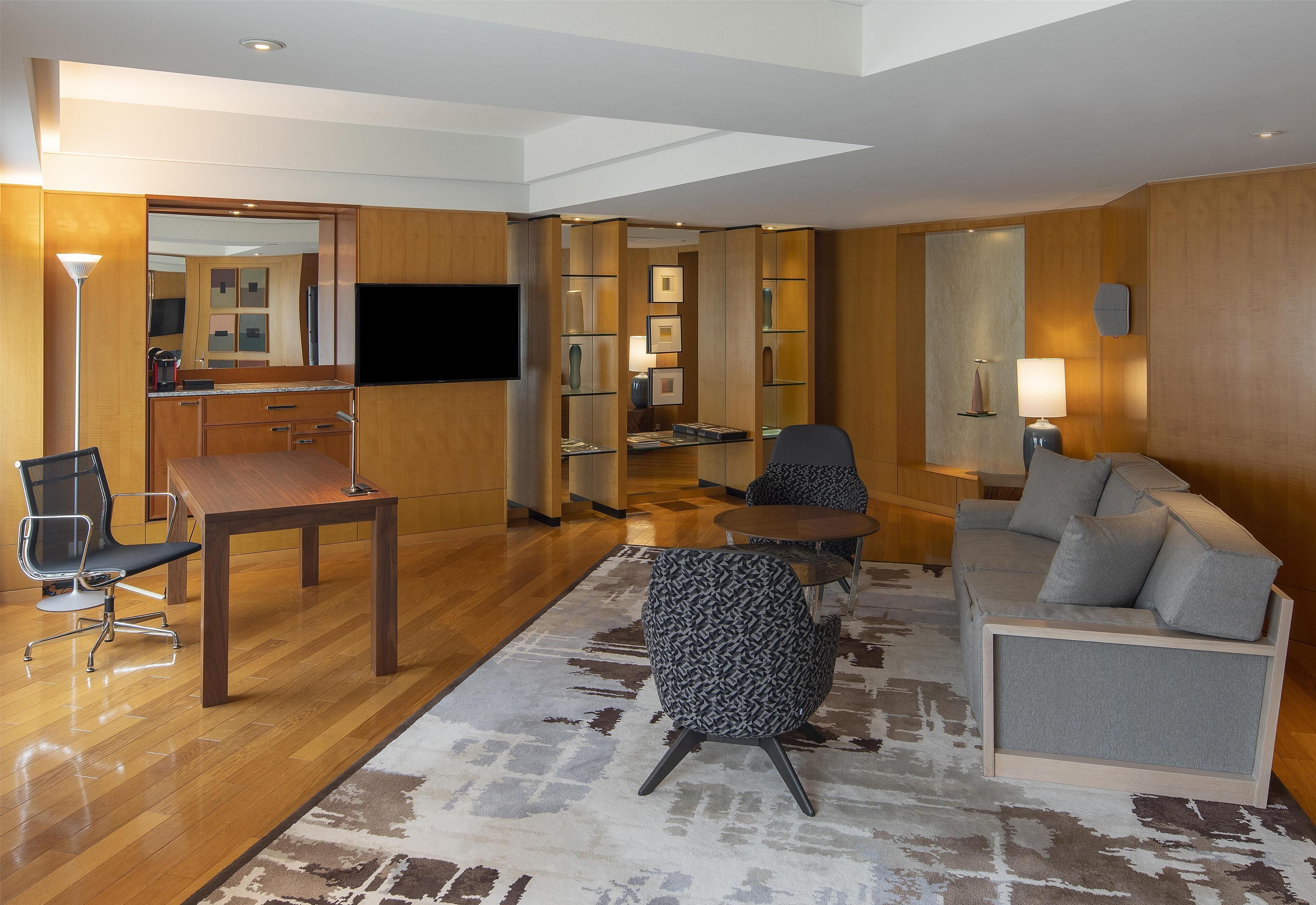 Una habitación de hotel moderna con suelo de madera, un sofá, una silla, una mesita, un televisor y un espejo.