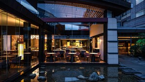 Un área de comedor al aire libre con mesas y sillas, una fuente de agua y una fachada de edificio moderna.