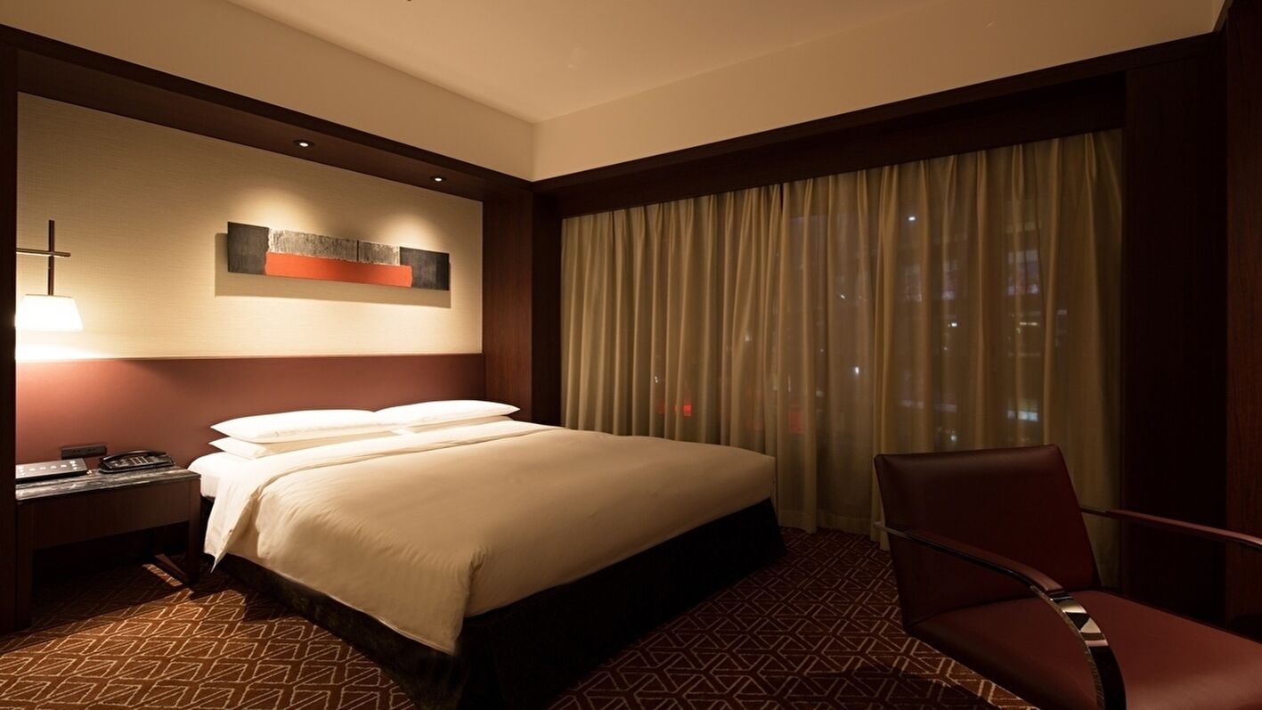 A hotel room with a large bed, a desk with a lamp, a chair, and a window with curtains.