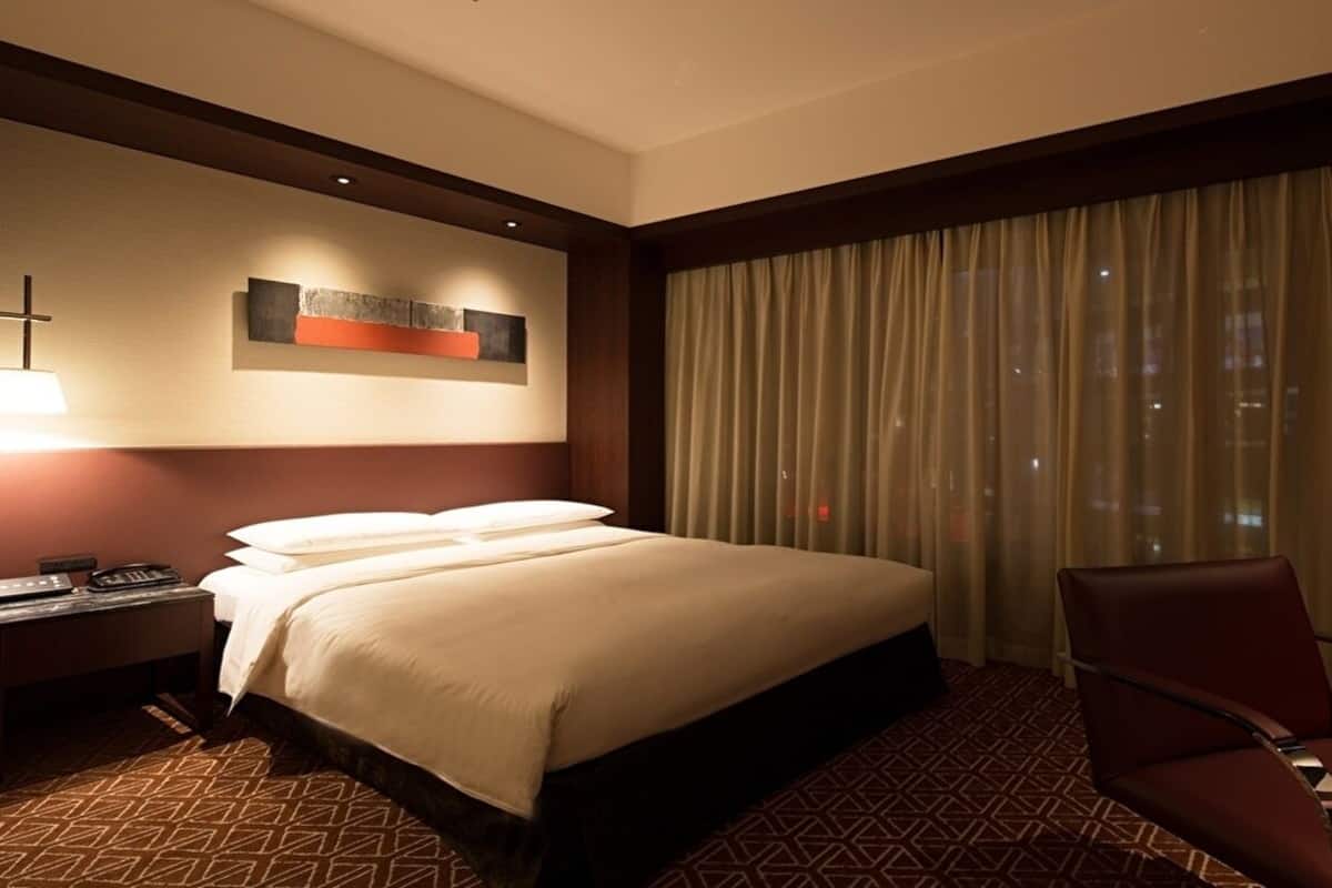 A hotel room with a large bed, a desk with a lamp, a chair, and a window with curtains.