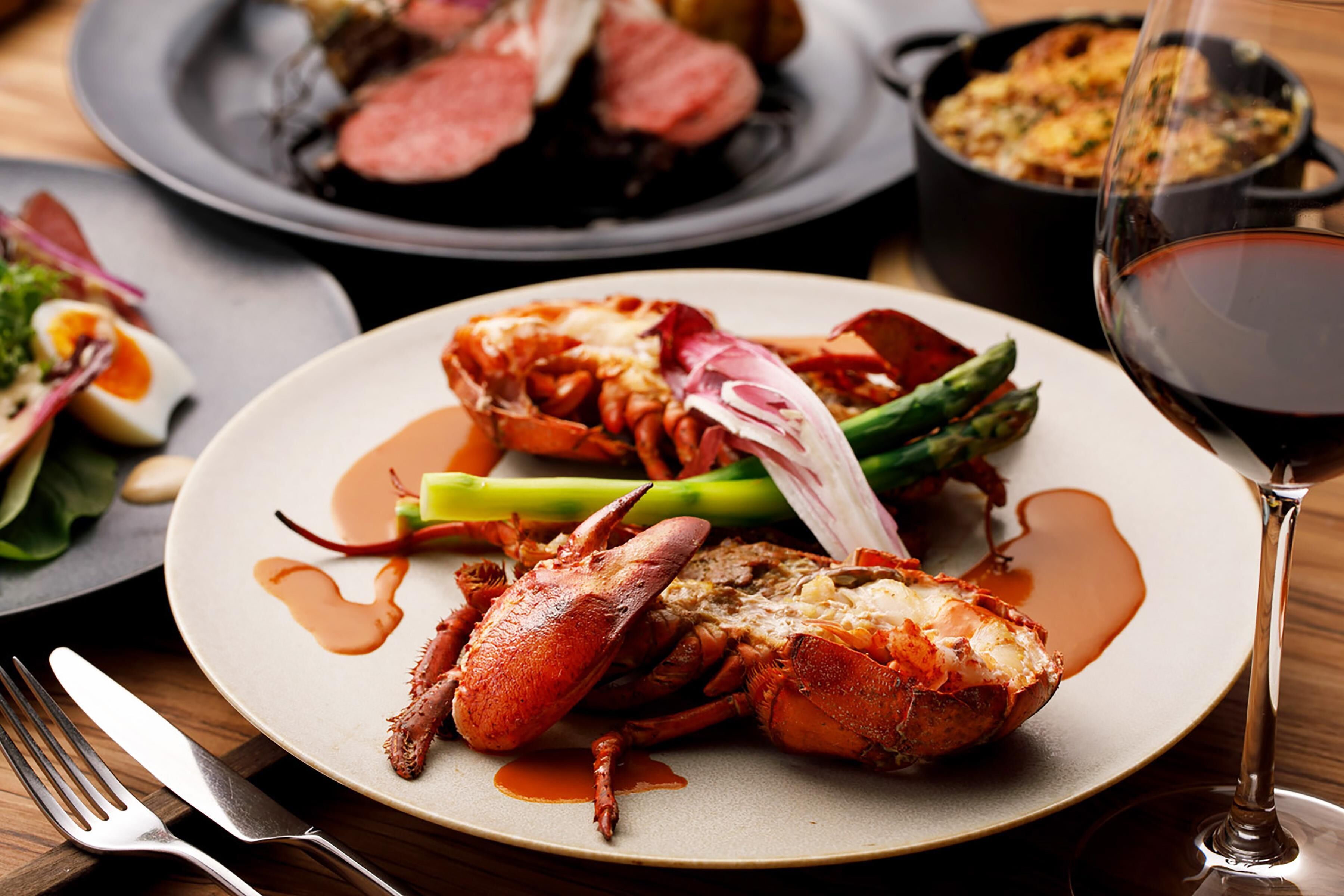 A lobster dish on a plate with asparagus and sauce, a fork, and a glass of red wine.