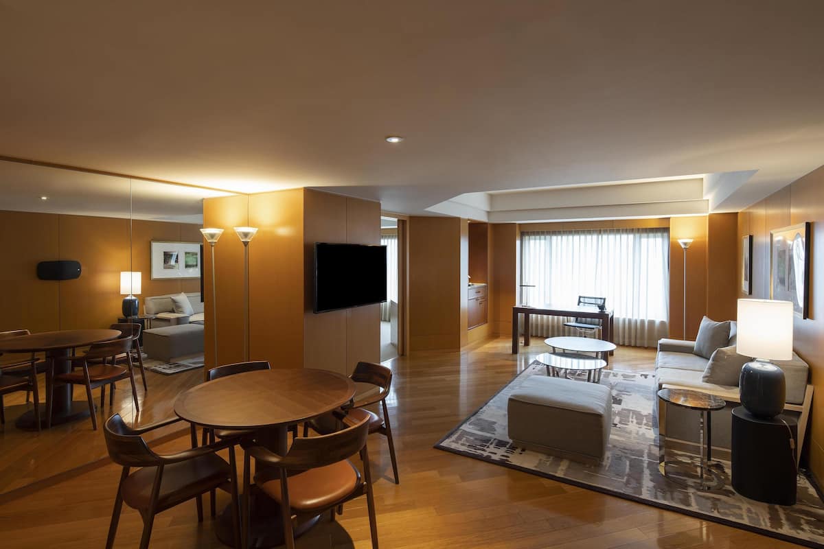 Ein modernes Hotelzimmer mit Essbereich, einem Wohnbereich mit Sofa und Fernseher sowie einem großen Fenster mit Vorhängen.