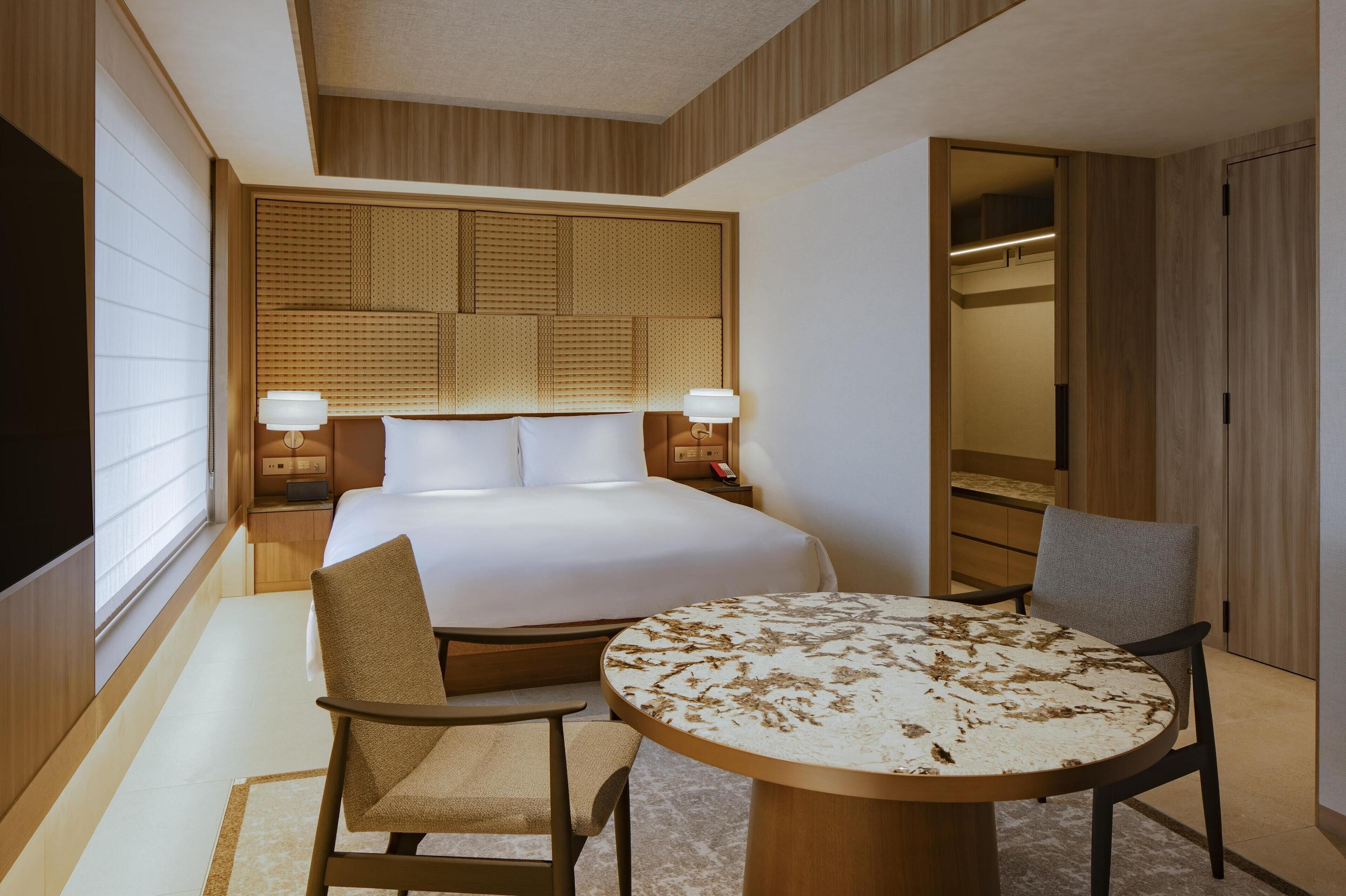Quarto de hotel com uma cama grande, duas mesas de cabeceira com abajures, uma mesa de jantar redonda e uma cadeira.