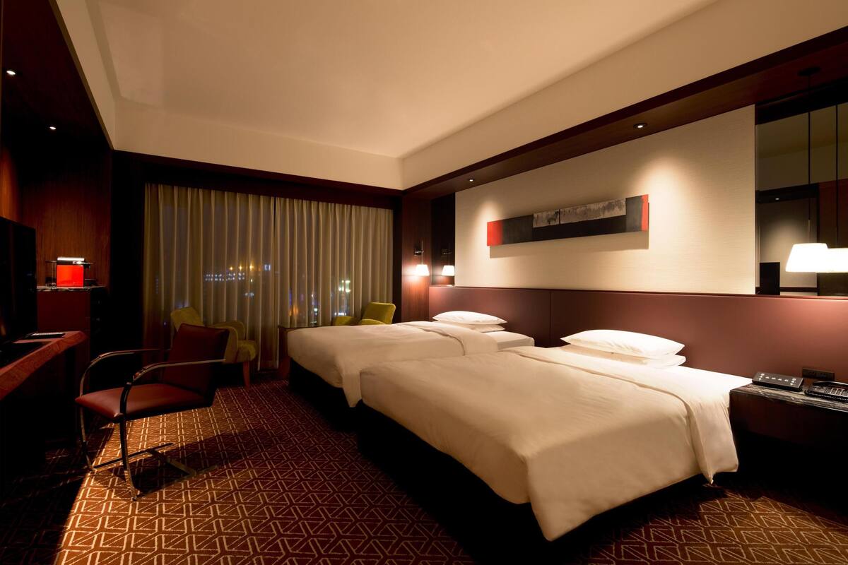 Ein Hotelzimmer mit zwei Betten, einem Schreibtisch, einem Stuhl, einem Fernseher und einem Fenster mit Vorhängen.