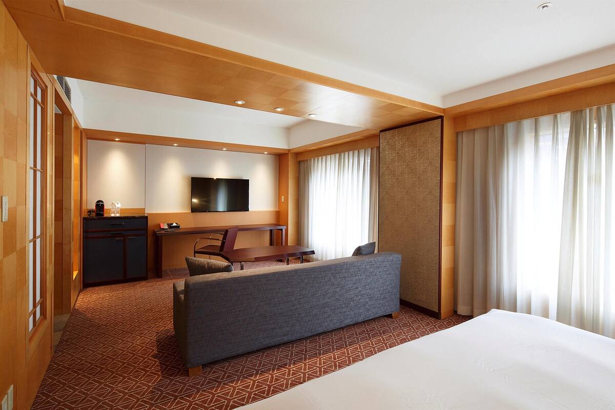Ein Hotelzimmer mit einem großen Bett, einer Sitzecke, einem Schreibtisch, einem Fernseher und einem Fenster mit Vorhängen.