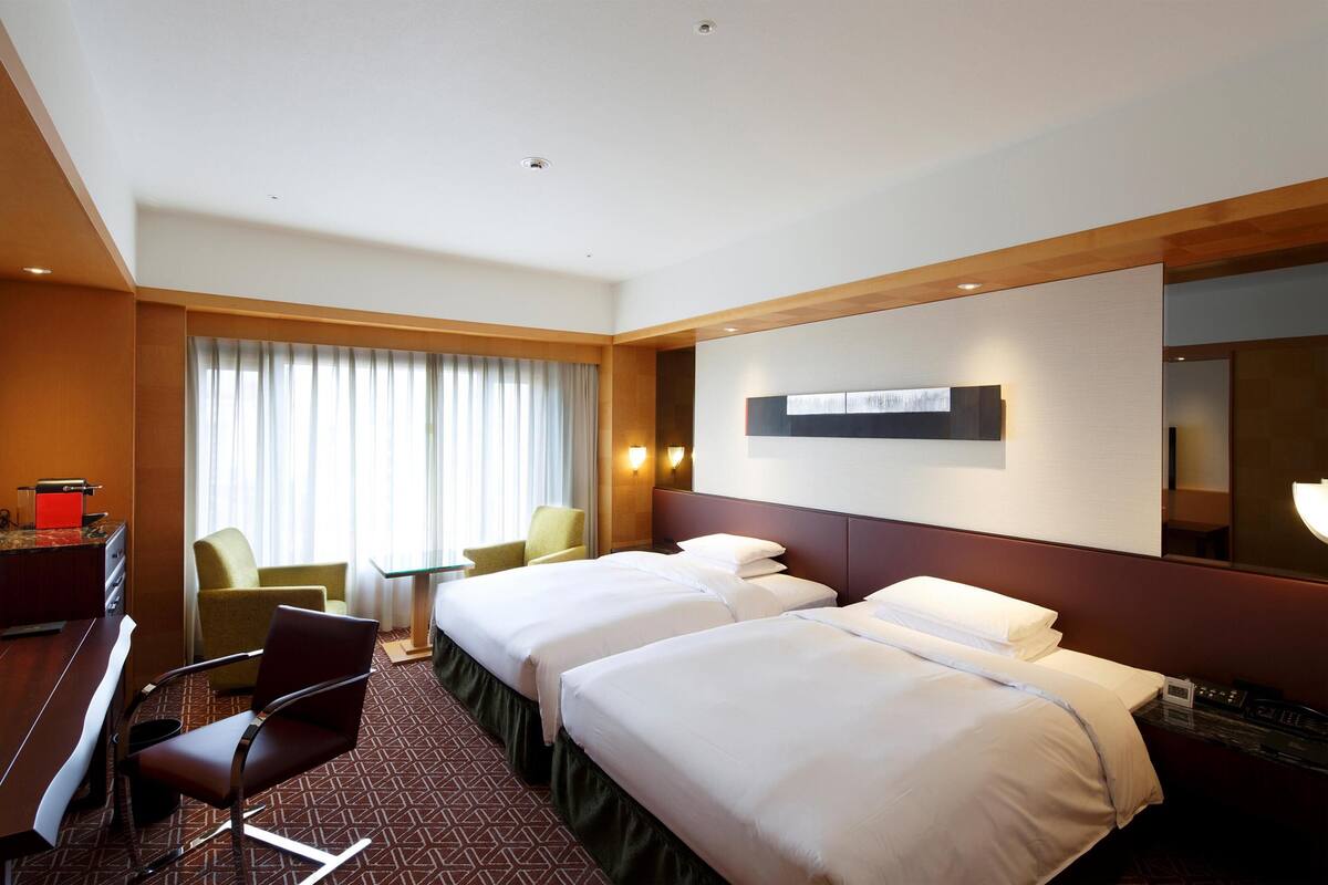 Ein Hotelzimmer mit zwei Betten, einem Schreibtisch, einem Sessel und einem großen Fenster mit Vorhängen.