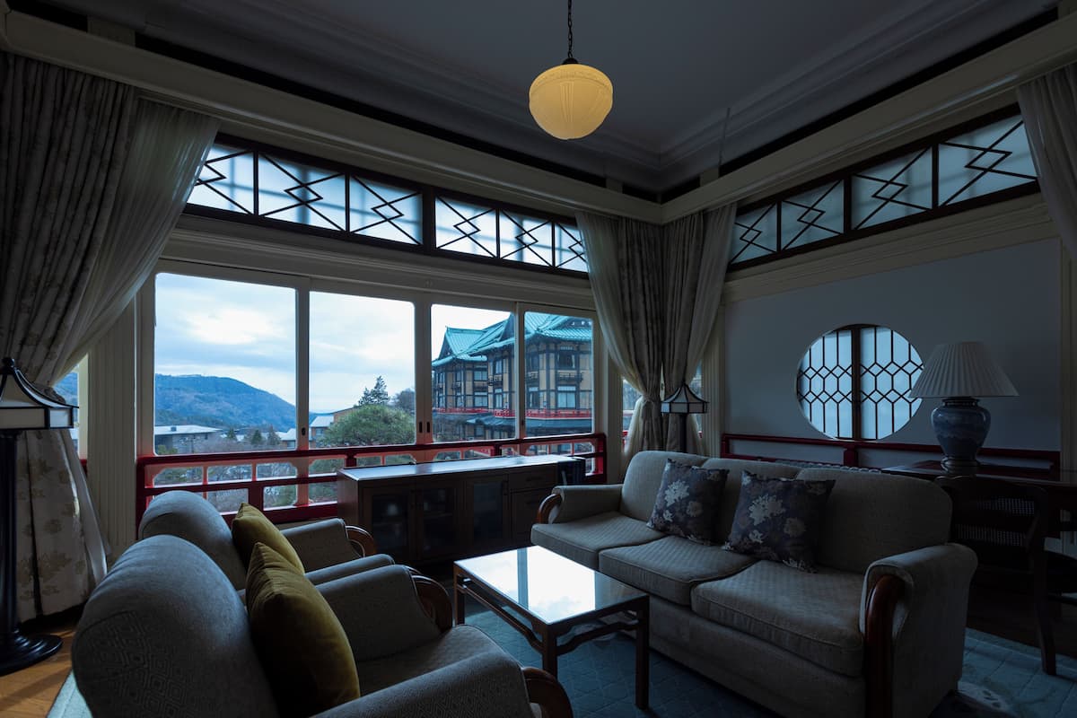 Un salón amplio con sofá, sillones y una mesa de centro. Ventanas grandes ofrecen vistas a un edificio y montañas.