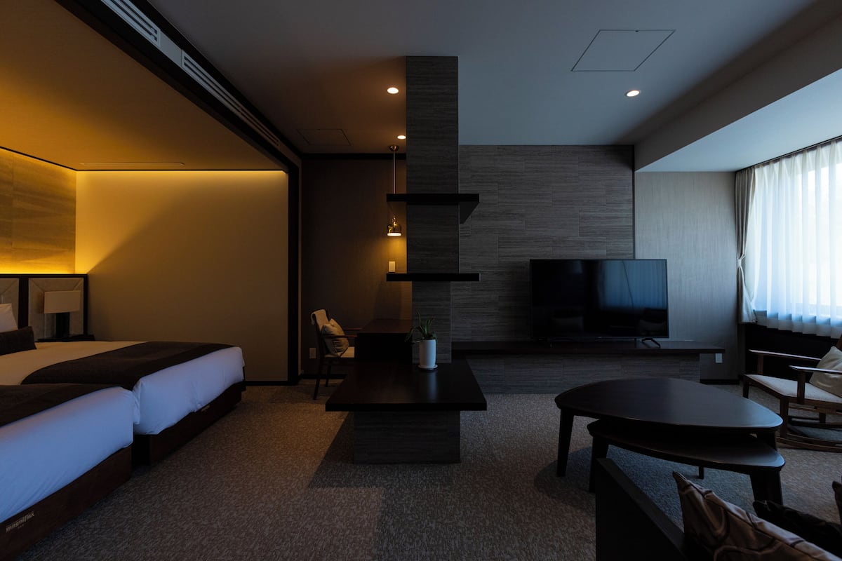 Una habitación de hotel moderna con una cama grande, un televisor de pantalla plana, una mesa de centro y una zona de estar con un sofá y sillones.