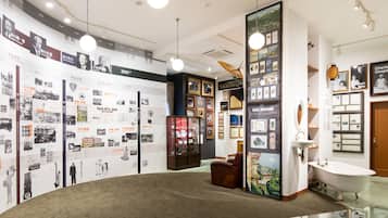 A hotel lobby with a round carpet, wall-mounted photo displays, a vintage bathtub, and a glass table with a book.
