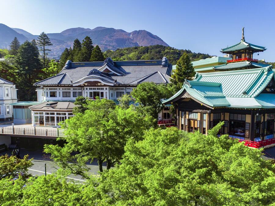 Een traditioneel Japans gebouw met een opvallend pagodatype dak, omgeven door weelderige begroeiing en een bergachtige achtergrond.