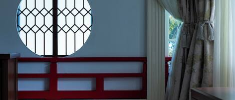Una habitación con un amplio ventanal circular con diseño geométrico, un banco rojo y blanco, y una cortina con flecos.