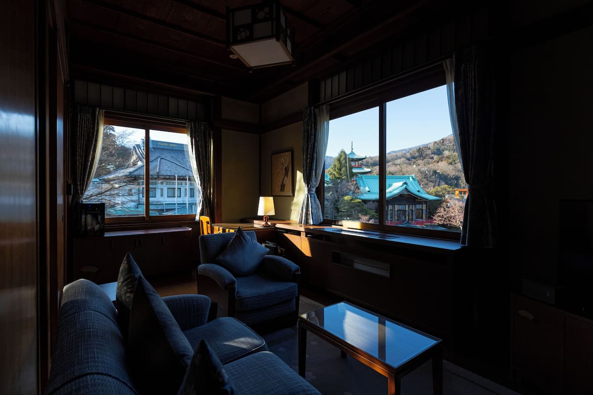 Una habitación con sofá, sillones y una mesa. Amplias ventanas con cortinas ofrecen vistas de un edificio tradicional.
