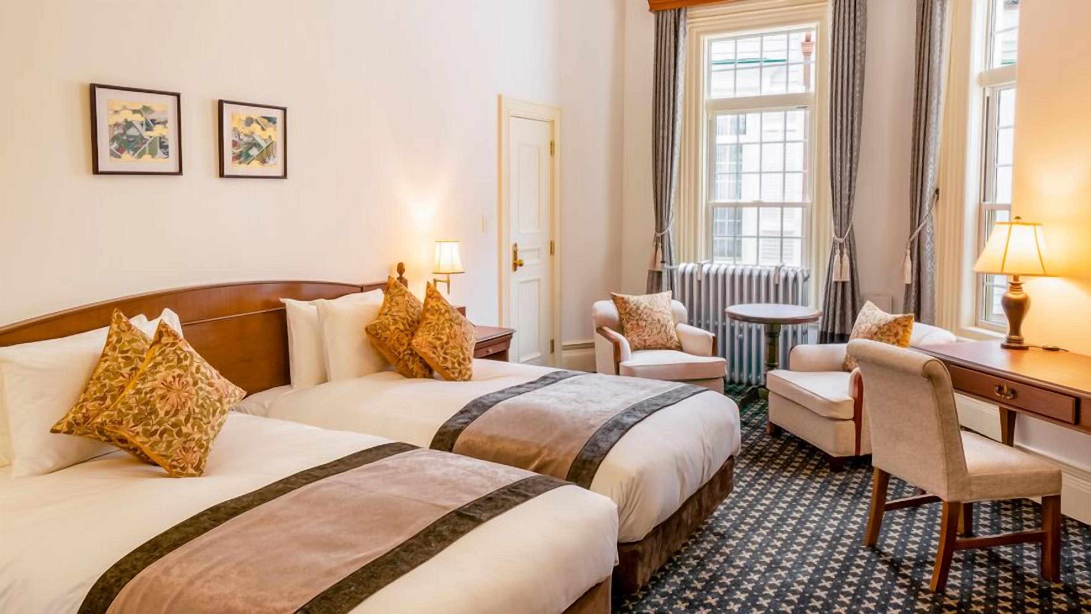 A hotel room with two beds, a desk, a chair, a table, and two framed pictures on the wall.