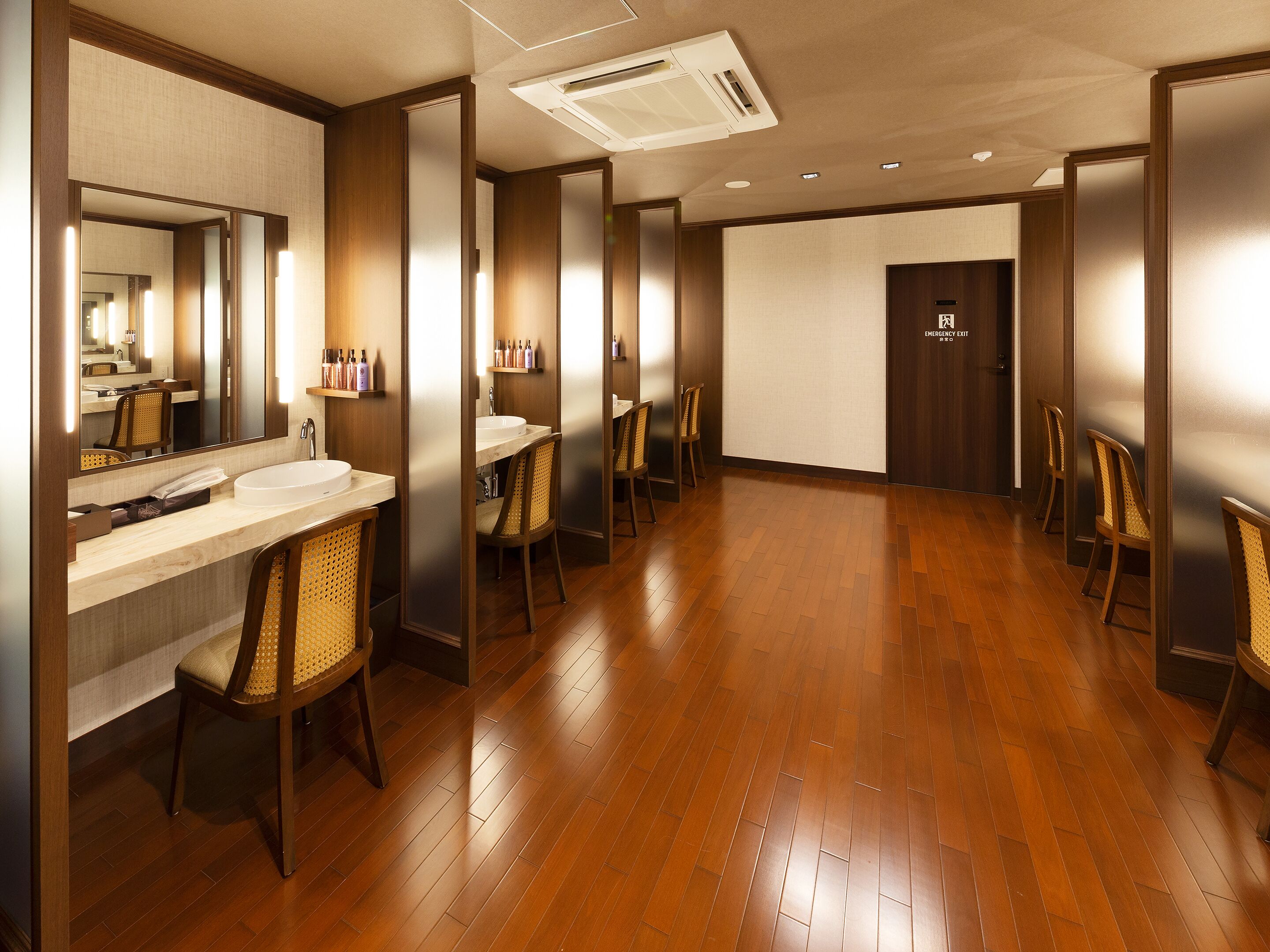A hotel bathroom with wooden flooring, multiple sinks, and chairs.