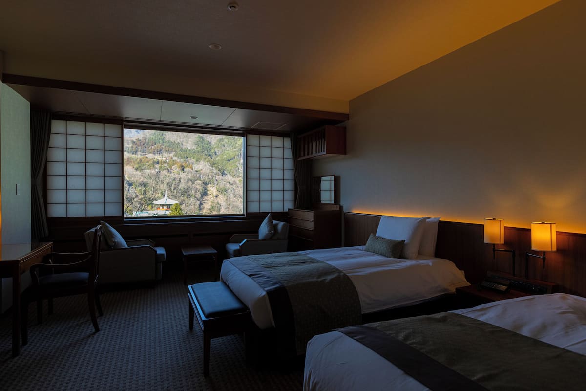 Habitación de hotel con dos camas, un escritorio y una silla. Se ve un ventanal con vistas a árboles y colinas.