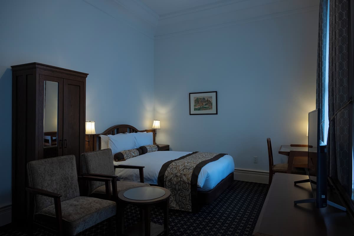 Una habitación de hotel con una cama, dos sillones, una mesita, un escritorio, un armario y un cuadro en la pared.