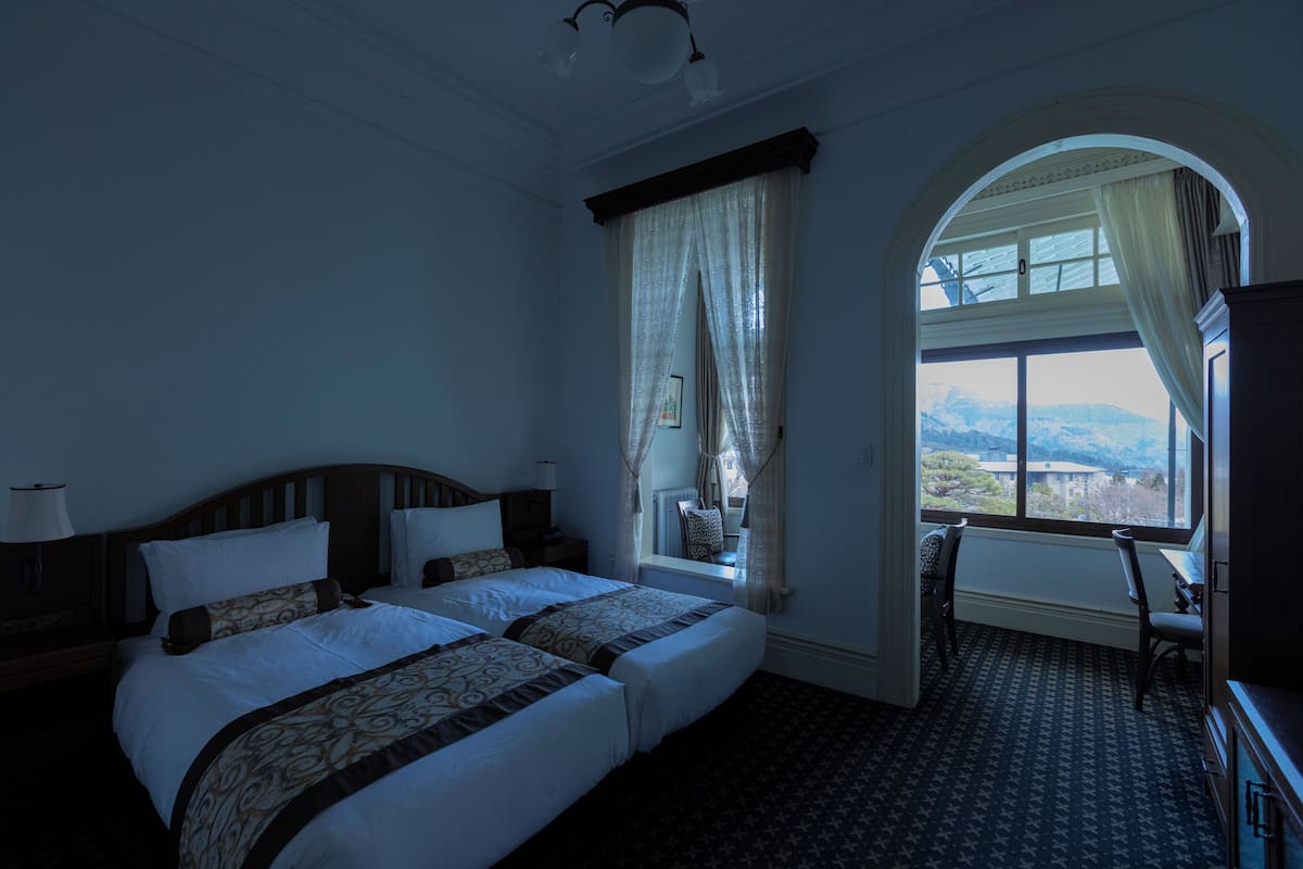 Habitación de hotel con dos camas, un escritorio y vistas a la montaña a través de un ventanal arqueado.