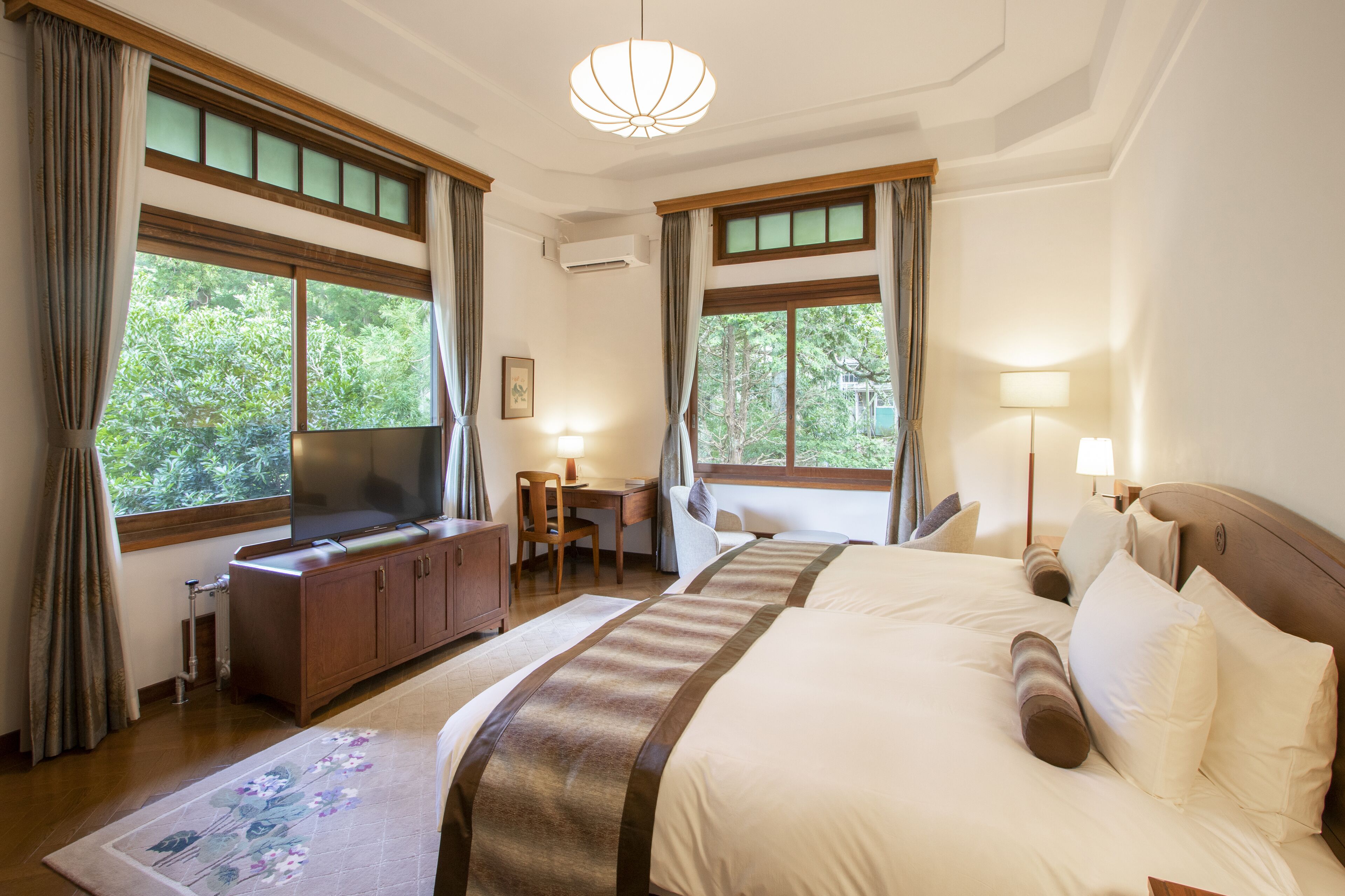 A hotel room with a large bed, a desk with a chair, a TV, and large windows with curtains.