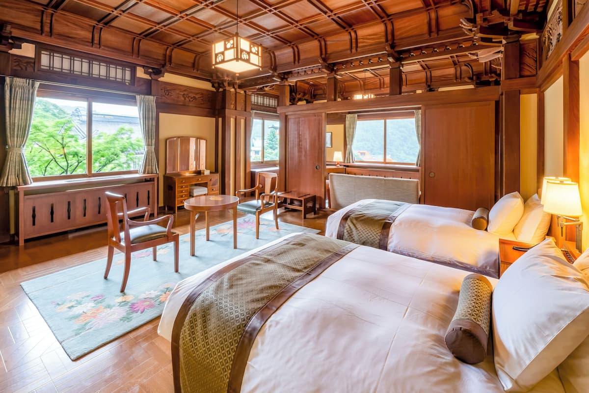 Habitación de hotel tradicional al estilo japonés, con vigas de madera, suelo de tatami, un escritorio, dos camas y grandes ventanales con vistas a zonas verdes.