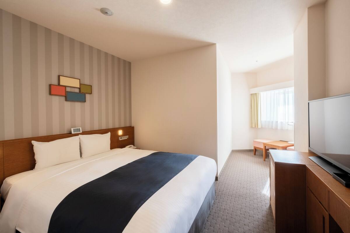 A hotel room with a large bed, a wooden headboard, a flat-screen TV, a bedside table, and a small desk with a chair.