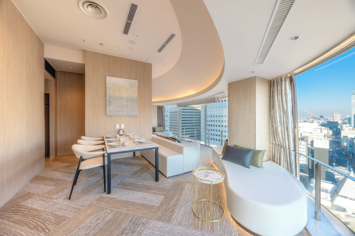 A modern hotel room with a dining area, a sofa, and a large window with a city view.