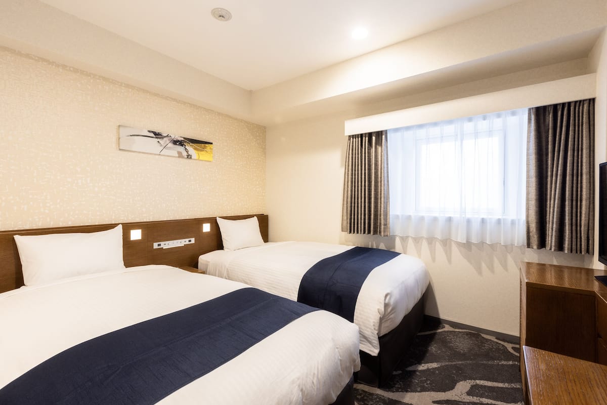A hotel room with two beds, a wooden headboard, a painting on the wall, a window with curtains, and a desk with a lamp.