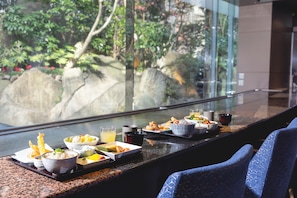A dining table with various dishes and drinks, set against a backdrop of a glass window overlooking a garden with rocks and greenery. - Shinjuku Washington Hotel Main (Tokyo)