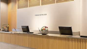 A reception desk labeled "FOREIGN VISITORS DESK" with three laptops, a pen, and a decorative vase with flowers.