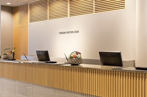 A reception desk labeled "FOREIGN VISITORS DESK" with three laptops, a pen, and a decorative vase with flowers.