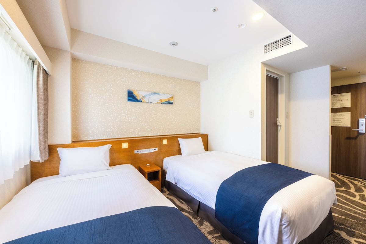 A hotel room with two beds, a small bedside table, a wall-mounted artwork, and a door leading to another room.