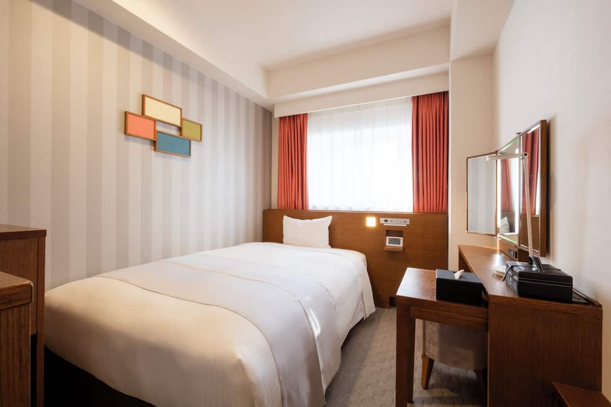 A hotel room with a large bed, a wooden desk, a mirror, and a window with curtains.