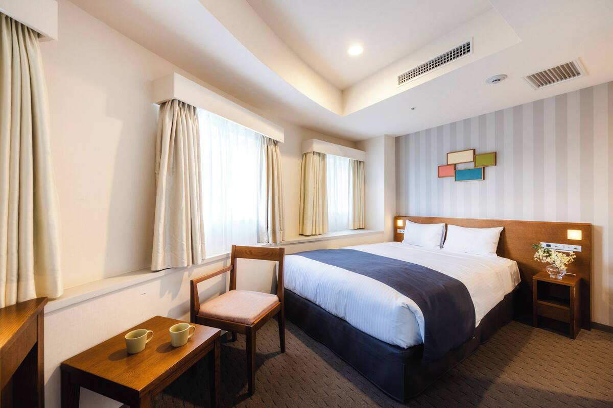 A hotel room with a bed, a chair, a small table with two cups, and a nightstand with a lamp.