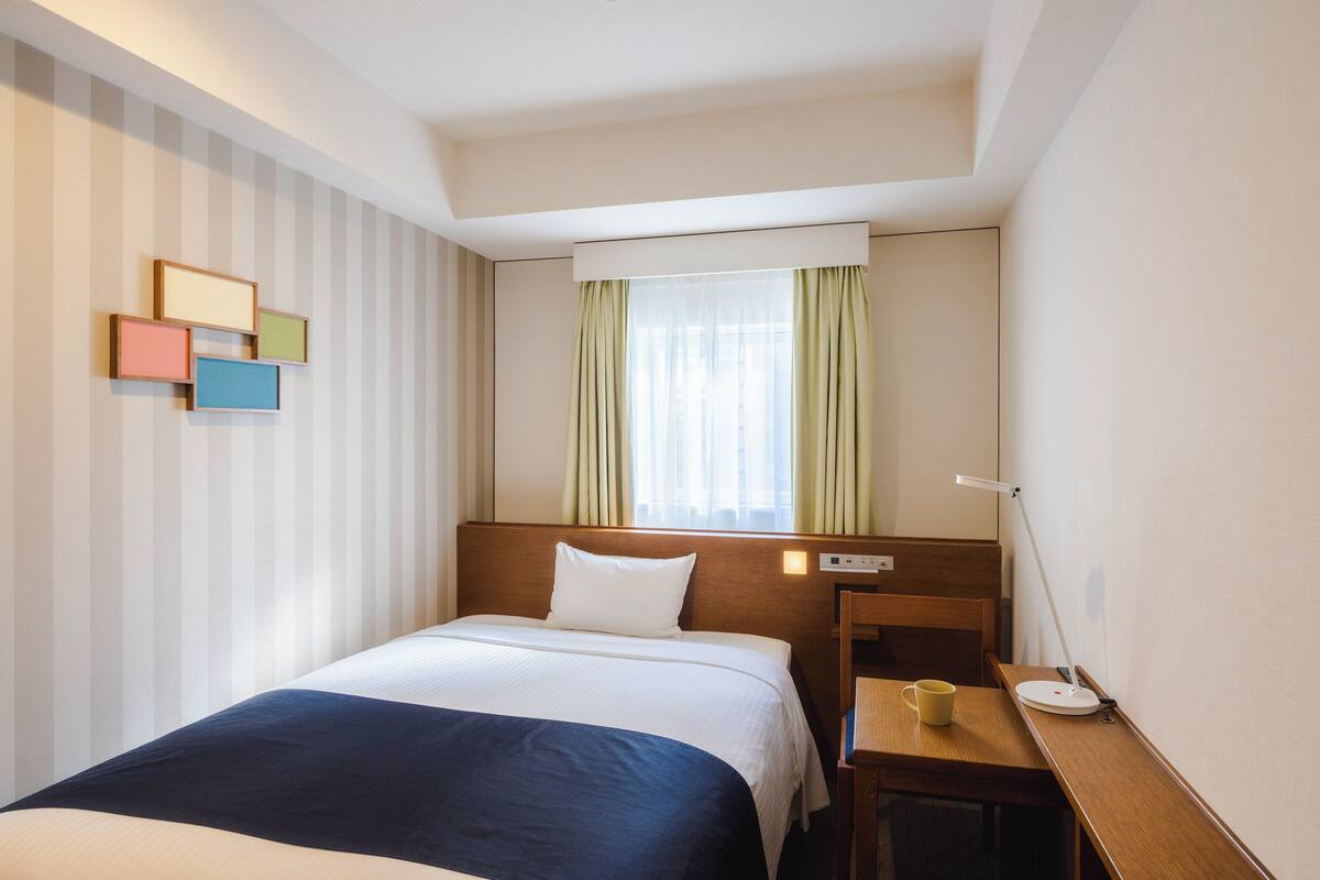 A hotel room with a bed, a desk, a chair, a lamp, and a window with curtains.