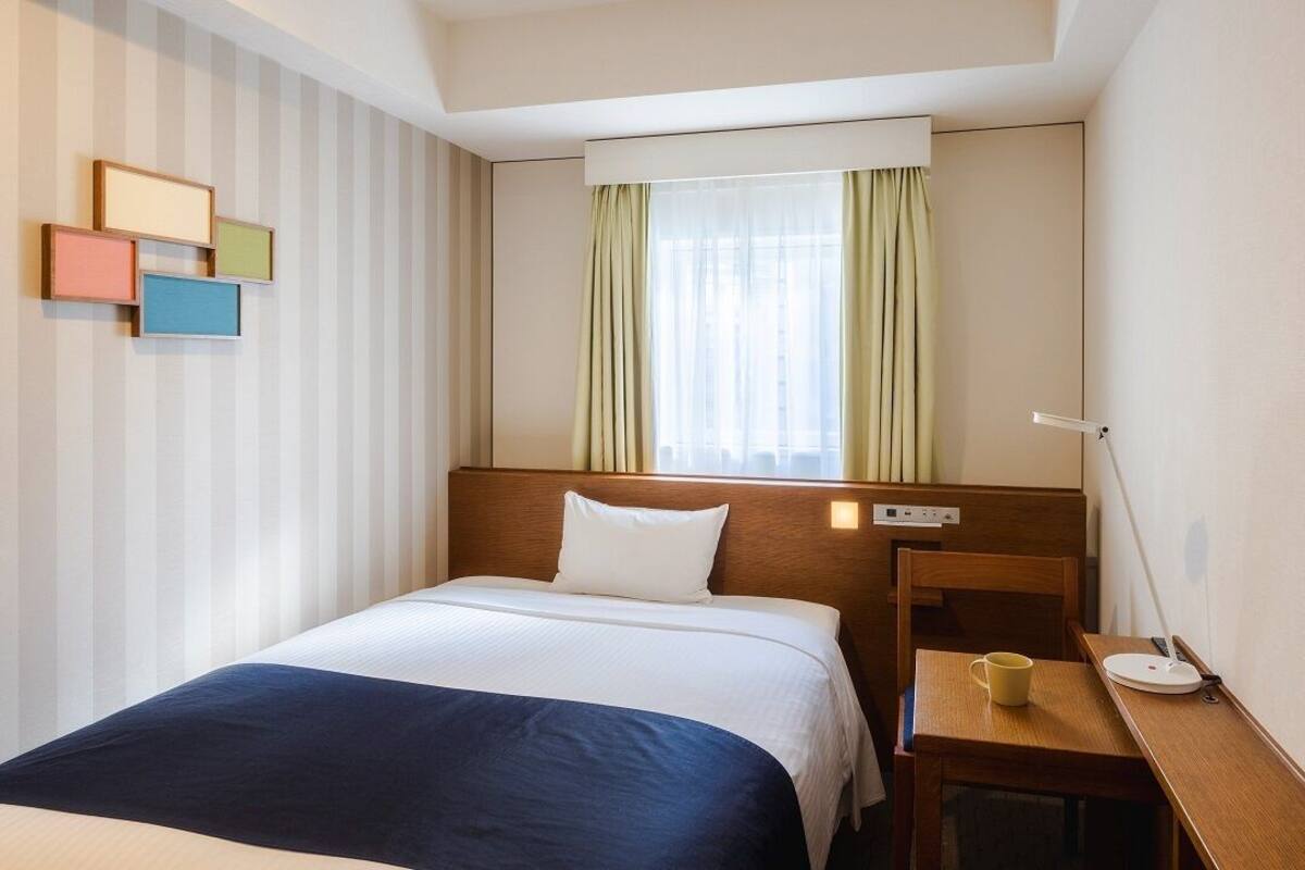A hotel room with a bed, bedside table, lamp, and a window with curtains.