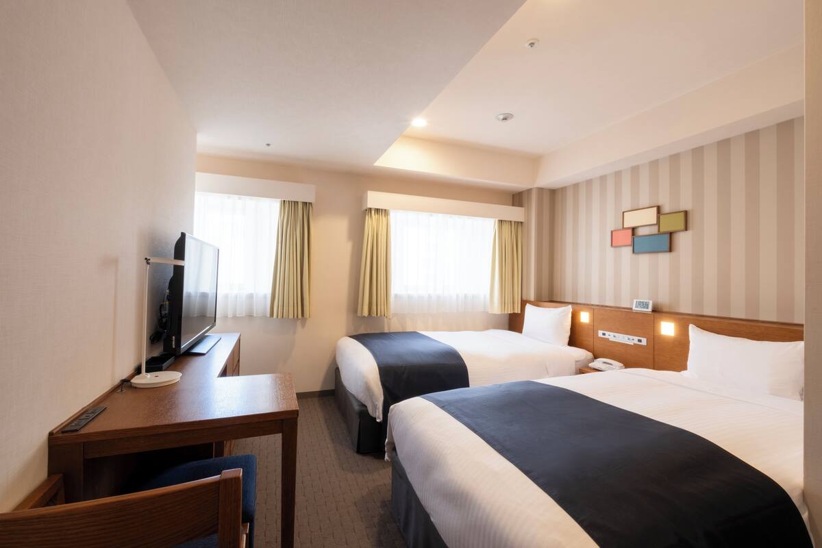 A hotel room with two beds, a desk, a TV, and a window with curtains.