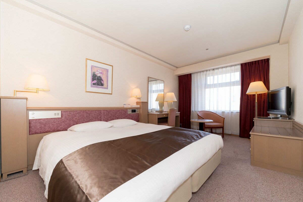 Una camera d'albergo con un letto grande, una scrivania, una sedia, una televisione e una finestra con tende.