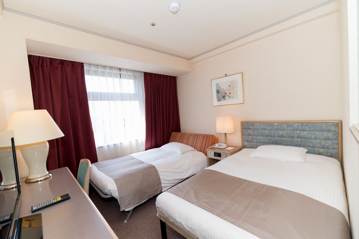 Una camera d'albergo con due letti, una scrivania con lampada, una sedia, un comodino con telefono e un quadro appeso al muro.
