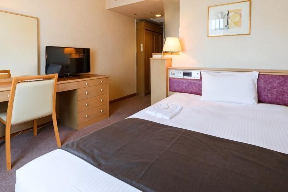Una camera d'albergo con un letto, una scrivania con televisore, una sedia, un comodino con lampada e una porta che dà su un bagno.