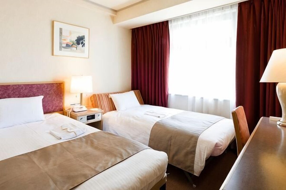 Una camera d'hotel con due letti, un comodino con lampada, una scrivania e una finestra con tende.