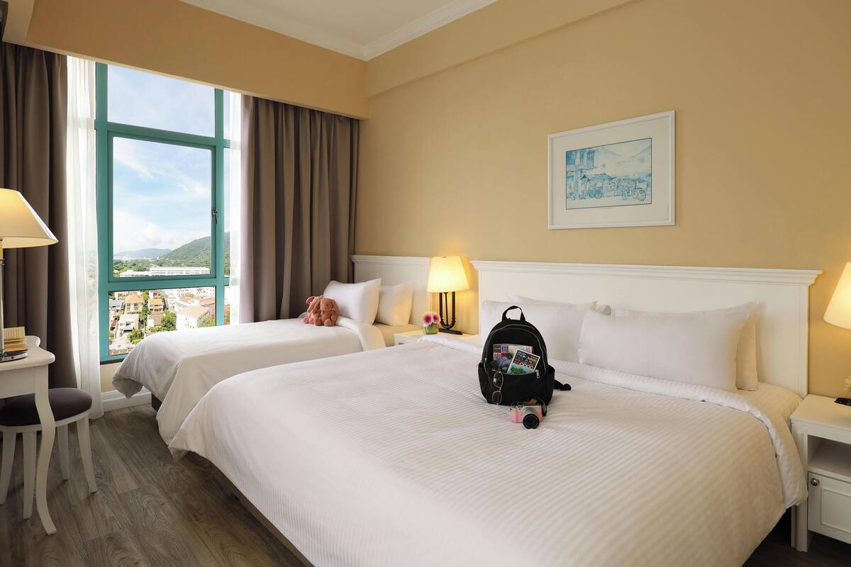 A hotel room with two beds, a large window with curtains, a desk with a lamp, and a framed picture on the wall.