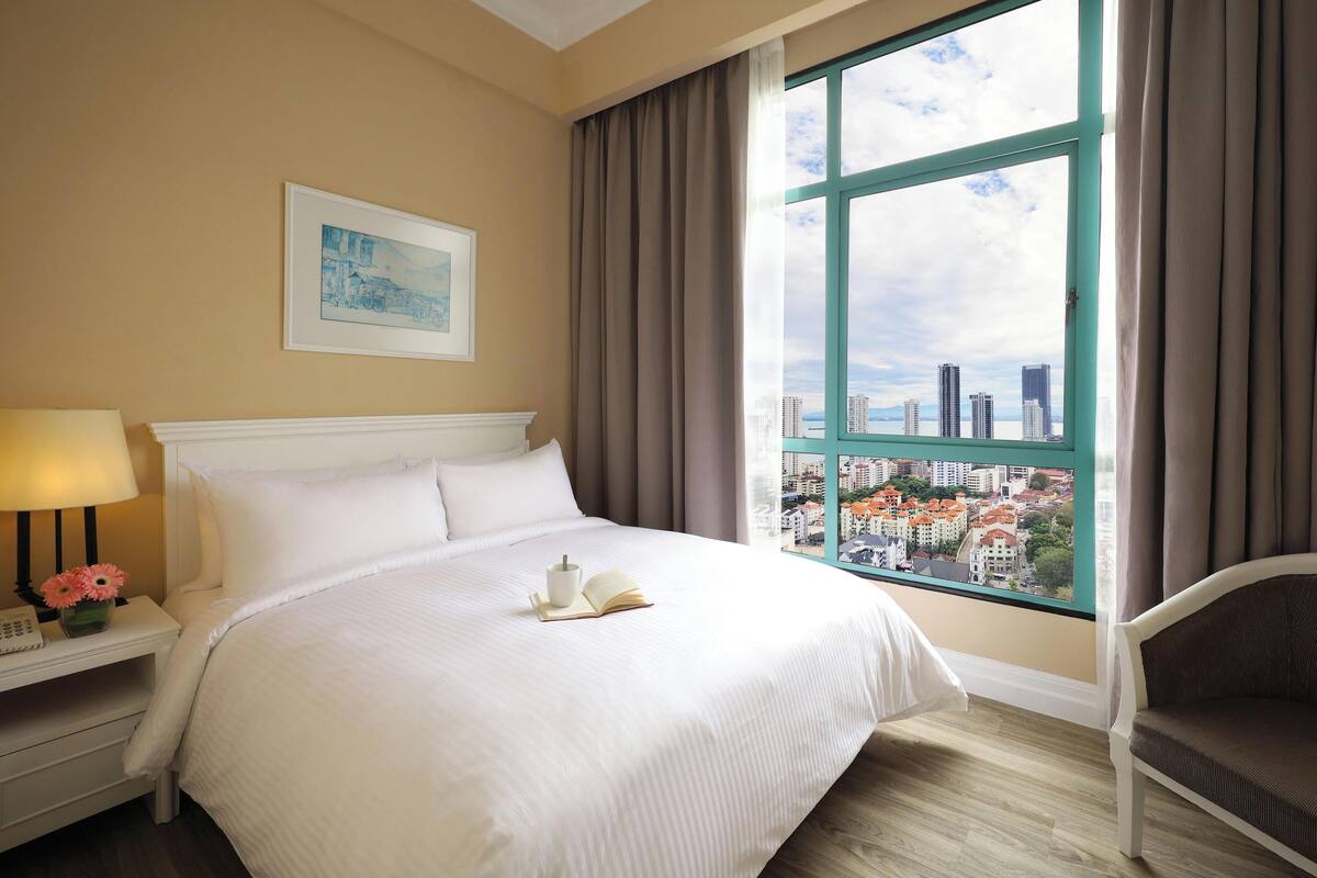 A hotel room with a large bed, a bedside table with a lamp and flowers, a chair, and a window with a city view.