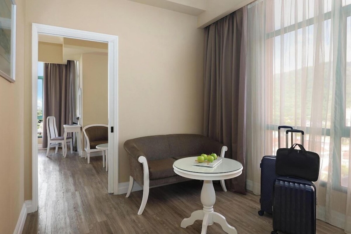 A hotel room with a sofa, a small round table with fruit, a chair, a suitcase, and a view through sheer curtains.