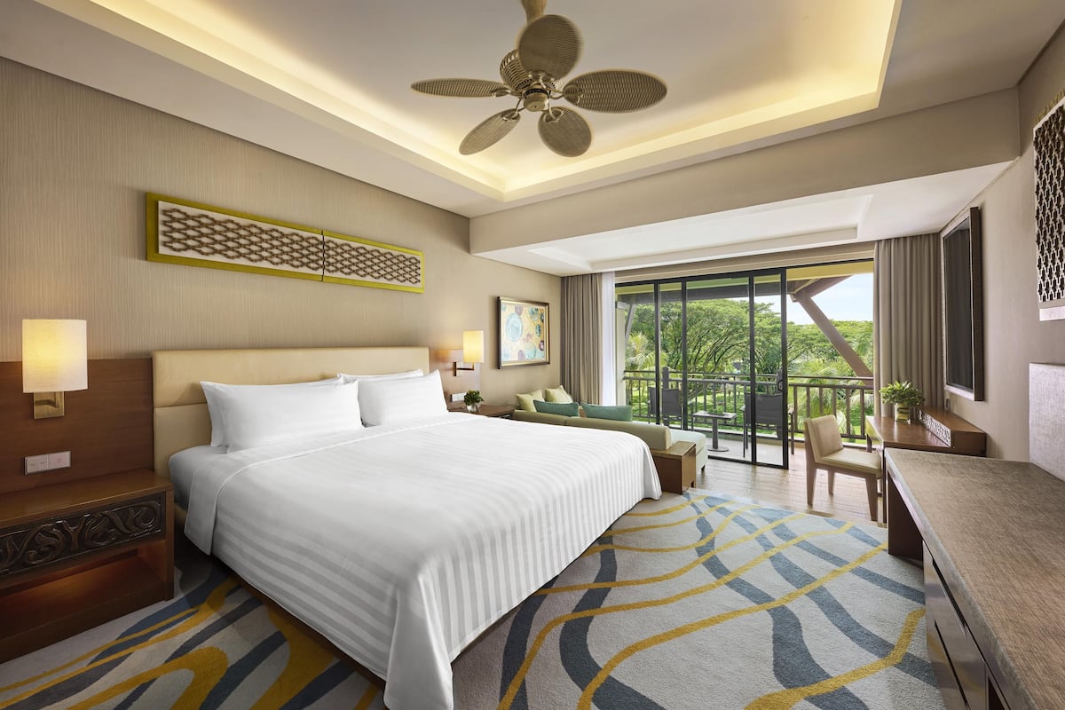 A hotel room with a large bed, a desk, a chair, a balcony with a view, and a ceiling fan.