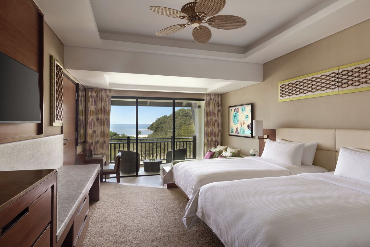 A hotel room with a balcony, a desk, two beds, and a ceiling fan.